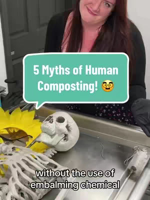 The image is a selfie-style shot featuring a woman in the background and a staged scene in the foreground. A turquoise speech bubble with the text "5 Myths of Human Composting!" frames the subject matter of the video while a stylized skull and skeleton are visible on a metallic surface. The woman has red hair and is wearing a dark top with a floral pattern. A yellow flower rests beside the skeleton in the foreground of the image. The image has a neutral, informative overall composition with a slightly comedic tone.