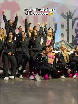 A dynamic photograph captures a group of women standing in a gym setting. They are all dressed in black athletic wear, many with their arms raised in a gesture of celebration or excitement. The background features a Gymshark logo and a classic car. The image has a lively atmosphere.