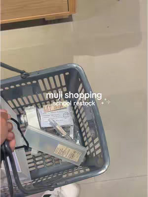 The image shows a shopping basket filled with Muji stationery supplies, with text overlays indicating 'muji shopping' and 'school restock'. The basket is gray and held by a person's hand wearing a white shoe, with a blurred background.