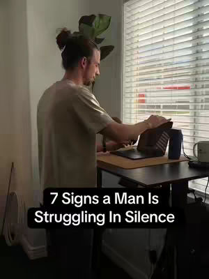 A man in a casual outfit stands near a window with blinds, working on a laptop at a desk. Text overlay highlights the video's topic: '7 Signs a Man Is Struggling In Silence'.