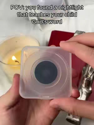 A hand is holding a small, translucent square object with a dark circle in the center, possibly a nightlight. Behind it is a lit candle, a deep red object, and part of a silver object. The background is blurred with soft lighting.