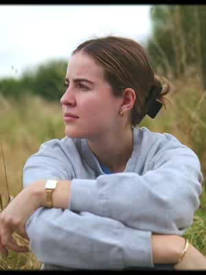A young woman with light skin, wearing a gray sweatshirt, is sitting in tall dry grass, looking off to the side with a thoughtful expression. Her arms are crossed, and she has a gold watch and bracelet. Her hair is pulled back in a bun, and she has a light background including trees and the sky visible.