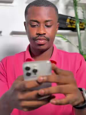 A man with dark hair and a mustache looks down at his phone, holding it in front of him. He wears a pink polo shirt. The setting seems to be indoors, with a blurred background.