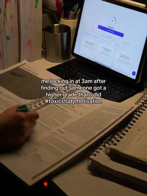 A well-lit shot of a desk setup with study materials. A hand with a pen interacts with open papers, while a laptop showing an interface sits to the right. A metallic mug and other stationery items are visible. The overall scene gives a sense of late-night studying or working.