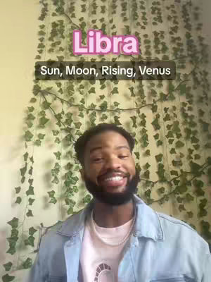 A person with a beard is smiling at the camera. They are wearing a light blue denim jacket over a light pink shirt. The background is covered in green vines.
