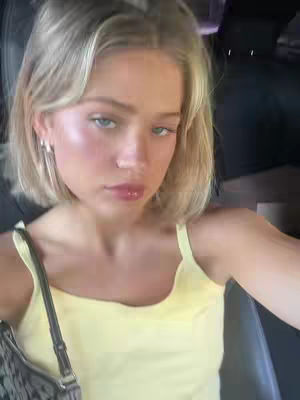 A selfie taken inside a car. The person in the image is a young woman with fair skin and blonde hair, styled in a short bob. She has light blue eyes and is wearing light pink lipstick and some light pink blush. She is wearing a light yellow tank top and has a black purse strap visible. The background is blurred with a dark interior and what appears to be a rearview mirror.