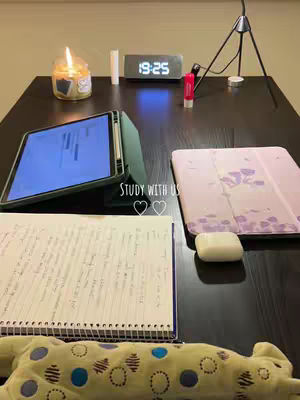 A top-down shot of a desk setup for studying. It features a dark brown table with a tablet and a notebook open, a lit candle, a digital clock, a tube of red lip gloss, and a pen. In the background, a small light is visible. The foreground includes a stuffed animal.