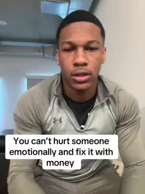 A young Black man is in the center of the frame, facing the camera and speaking. The background features a blurred room, possibly a home or office space. The man is wearing a gray Under Armour zip-up and a black shirt. The overlay text is in a white box at the bottom.