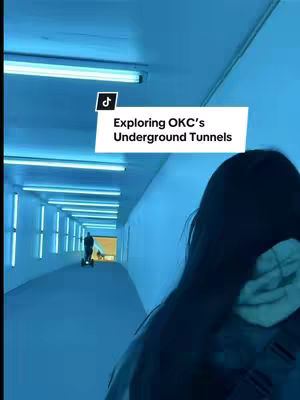 A person with long dark hair and a grey hoodie is in the foreground, facing towards the right. The rest of the image shows a long, blue-lit tunnel, with another person and a scooter in the distance. A white text box at the top left says, "Exploring OKC's Underground Tunnels."