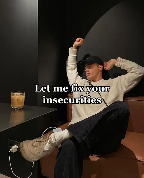 A young man is sitting in a dimly lit space, resting on a brown seat. He's wearing a white sweater, black cap, and dark-colored trousers, with white high socks and sneakers. He has his arms stretched above his head, giving the impression he is stretching. There's a glass of beverage on a small table next to him. Text overlayed says, 'Let me fix your insecurities'. The wall and the background are in dark colors.