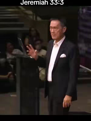 A middle-aged man in a black suit with a white dress shirt and pocket square stands at a lectern with a laptop on it, gesturing with his right hand. Behind him, a blurred audience and a dark background provide context. Text at the top indicates the Bible verse Jeremiah 33:3.