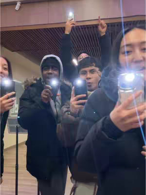 A group of young people, likely in a church or youth group setting, are holding up their phones with the flashlights on, creating a bright light effect. They appear to be interacting, one individual is in the foreground holding the camera close, smiling. Their expressions suggest joy and excitement, and the image is well-lit.