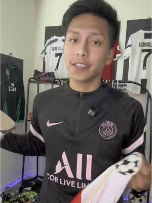 A young man stands in front of a wall with soccer jerseys, holding a jersey and soccer  balls, speaking directly to the camera. He wears a black sweatshirt and appears to be demonstrating how to fix a soccer jersey.