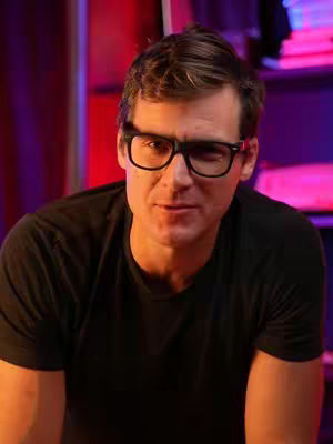A man with short brown hair and glasses is centered in the frame, looking directly at the camera with a slight smile. He's wearing a black t-shirt. The background is blurred but suggests a room with purple and red lighting.