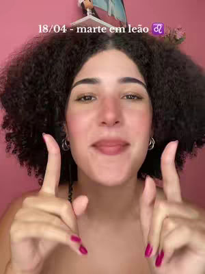 A woman with dark curly hair and fair skin is the primary focus of the image, set against a pink background. She is looking towards the viewer with a slight smile. Her fingers are extended upward forming a gesture. The text overlay reads "18/04 - marte em leão ♌" at the top of the frame.
