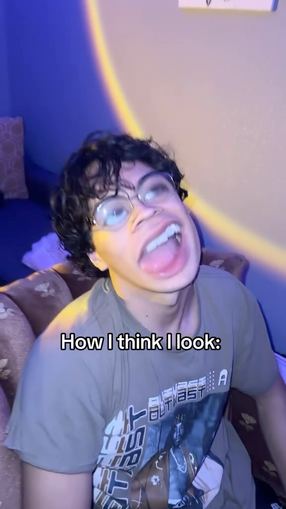 A young man with curly dark hair and glasses is captured in a close-up, making an exaggerated facial expression with his mouth wide open, showing his teeth and tongue. The background has a wall with a blue and yellow light gradient. The text overlay says 'How I think I look:' in a white, sans-serif font.