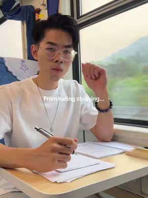 A young man with glasses writes in a notebook at a table on a train. The background shows a blurry landscape viewed through the train window. The scene is lit with natural light.