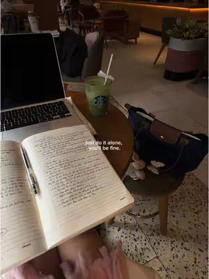 The image captures a relaxed, intimate moment in a cafe setting. A laptop sits open on a table, along with a handwritten journal and a pen, suggesting personal reflection or study. A green drink with a straw and a handbag add personal touches. The scene is bathed in warm, soft lighting, with a blurred background showing other tables and chairs. The overall composition conveys a sense of solitude and introspection.