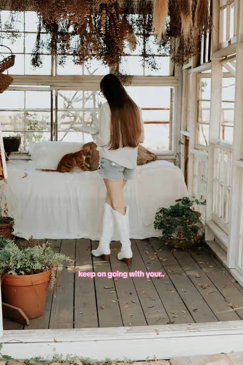 A woman with long brown hair stands in a sunlit room with a bed, a cat, and plants. She is wearing a white shirt and jean shorts with white boots. The walls and windows have a wooden frame and the bed has a white sheet.