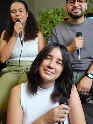 Three people with microphones are positioned in front of a blurred green background. A woman in the foreground is smiling, holding a microphone, with two other people behind holding mics. The overall composition is brightly lit, suggesting a performance or recording setting.