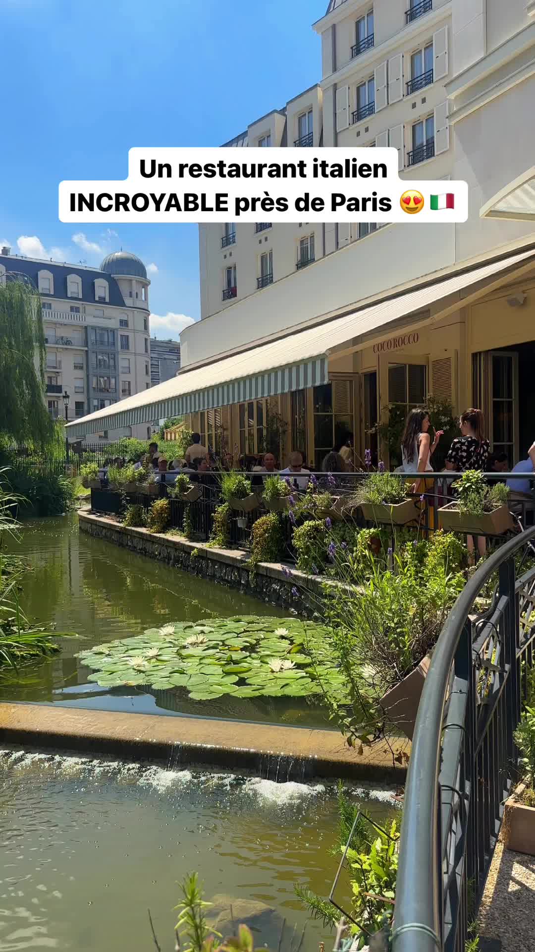 The cover image showcases an Italian restaurant scene. A waterway with lily pads and lush greenery is in the foreground, with a stone wall. The restaurant, featuring a white facade and striped awnings, extends along the water's edge. People are visible dining in the outdoor seating area, enjoying the summer atmosphere. The sky is blue in the background. Text overlays describe the restaurant.