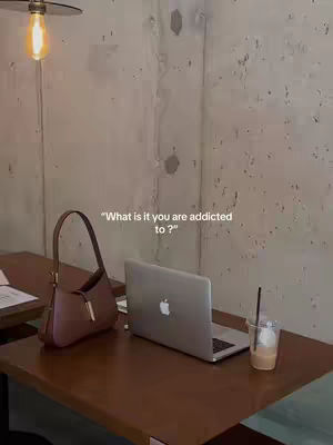 A well-lit image shows a cafe setting with a table set for work. A laptop is open on the table with a cup of iced coffee, a stylish purse, and some papers.