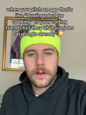 A man in a neon green beanie and black hoodie looks directly at the camera, presumably speaking. Text overlays the image describing the scenario: "when you pitch an app that's like Duolingo but for cooking to your coding friends but they all "have jobs at google already". The background is a frame with a painting.