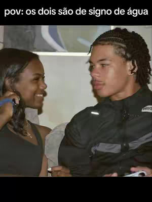 A photograph depicts a couple, a woman and a man are facing each other at a close range. The woman is on the left side of the image, with her dark hair pulled back. She is smiling while holding a blue object. The man is on the right side, with intricately braided hair. They appear to be in casual clothing. The background is blurred, soft-filtered.
