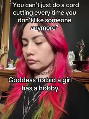 A woman with bright pink hair is centered in the frame, set against a cluttered background with dark tones. She wears a black top and multiple necklaces. Text overlays in black on white appear at the top and bottom, offering commentary on the image.