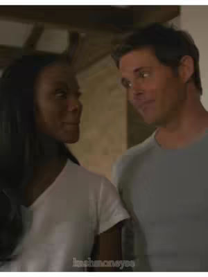 A still image from a movie featuring a woman and a man looking at each other. The woman has dark hair and is wearing a white t-shirt. The man has short hair and is wearing a light-blue t-shirt. They are in what seems to be an interior setting, possibly a home, with a neutral background. The image focuses on their faces and upper body.