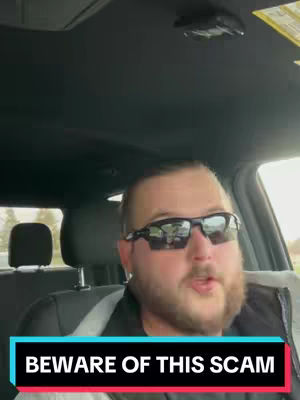 A man wearing sunglasses is filmed from the chest up, driving a car. The image highlights his upper torso and face, with the car interior and outside scenery blurred in the background.