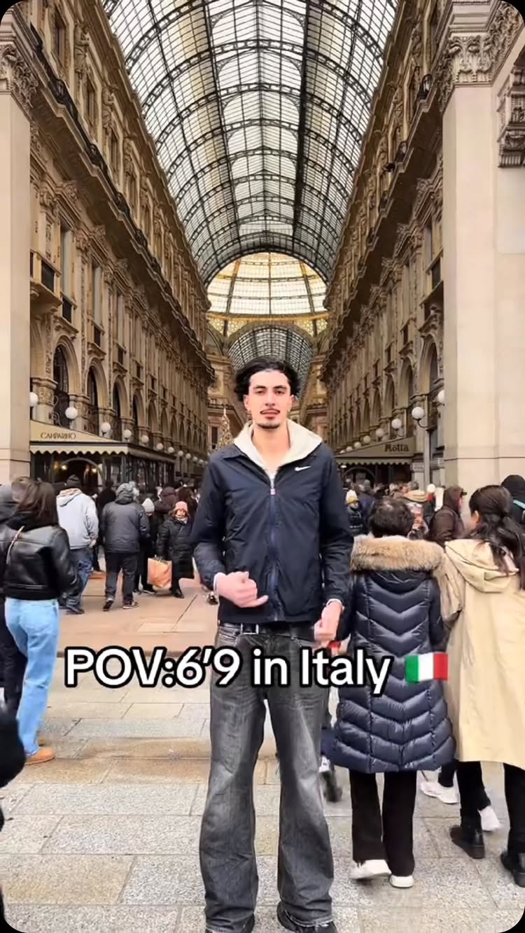 A man in a black jacket and jeans stands centered, with a historic building with glass roof in the background. Text overlayed says 'POV: 6'9 in Italy' with the Italian flag.