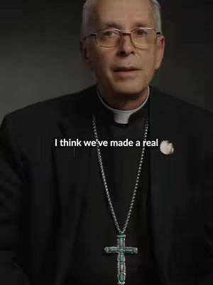 A medium shot features a man in a dark suit and clerical collar, wearing glasses, and a silver chain with a large cross. Text overlayed reads, "I think we've made a real". The background is subtly blurred.