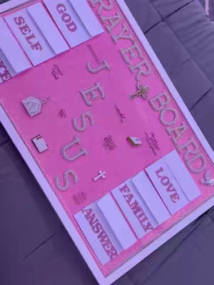 A pink prayer board on a bed, featuring the word 'JESUS' spelled out with glittering letters and other words like 'GOD,' 'SELF,' and 'LOVE.' The board has a white border and decorative accents.