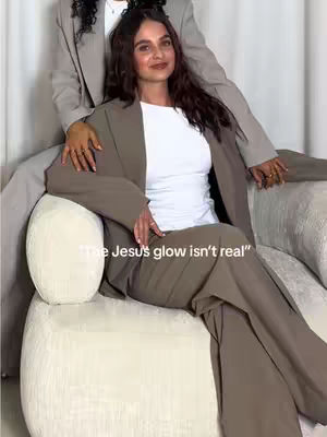 The image features a woman seated on a cream-colored, rounded-edge ottoman, dressed in a tan pantsuit and a white top. Another person, also in a tan suit, is partially visible behind her, with their arms resting on her shoulders. Text overlayed at the bottom reads, "The Jesus' glow isn't real."