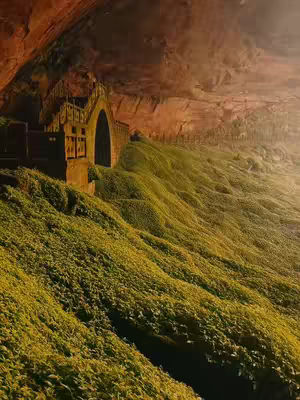 A landscape image featuring a stone structure nestled in a cave. Lush green vegetation covers a sloping hillside, leading up to the structure with an arched doorway. The scene is bathed in warm sunlight from an unseen source, creating strong shadows and highlighting textures. The cave's reddish-brown walls frame the vibrant green and golden hues, conveying a sense of serenity and mystery.