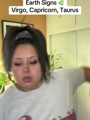 A woman with dark hair in a ponytail is looking at the camera with a slightly surprised or animated expression. She is wearing a white t-shirt. The background consists of a wall with a framed picture and part of a doorway or window. A text overlay at the top says Earth Signs and lists Virgo, Capricorn, and Taurus.