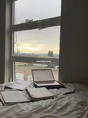 The image shows a well-lit bedroom scene with a bed covered in white bedding. A large window on the left reveals an overcast sky with a glimpse of a cityscape. On the bed, an open laptop and several notebooks sit near the window, alongside a pen, suggesting a study or work setup. The text 'evening study' is overlaid on the image.