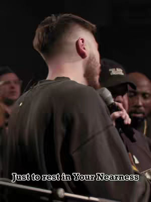 The cover image captures a man from behind, slightly angled towards the right. The man, who appears to be the central figure, is wearing a dark-colored long-sleeve shirt. His short hair has a distinct fade along the sides with the top styled, and he has a beard. A microphone is held near his face, suggesting he might be speaking or singing. Behind him, other people are visible out of focus, and the lighting suggests a lower-lit performance or gathering. Text at the bottom of the image reads, \"Just to rest in Your Nearness\".