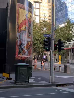 An outdoor shot captures an Australian city street with a large, curved pillar displaying a vibrant painting of a saint on horseback. People are walking on the sidewalk, and traffic signals are visible in the background, set against a backdrop of tall buildings and a bright sky. The image conveys a blend of urban life and religious symbolism.