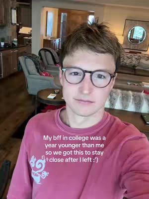 A young man with short brown hair and glasses smiles directly at the camera. He is wearing a pink long-sleeved shirt. The background is blurred, suggesting an interior setting with soft lighting and visible furniture.