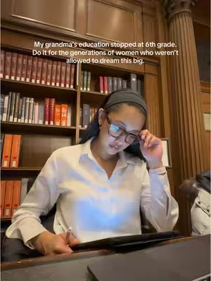 A young woman is seated at a desk in a library, her face illuminated by the light of a tablet. She wears glasses and a dark headband, and a bookcase filled with books forms the backdrop. The image is shot from a slightly low angle.