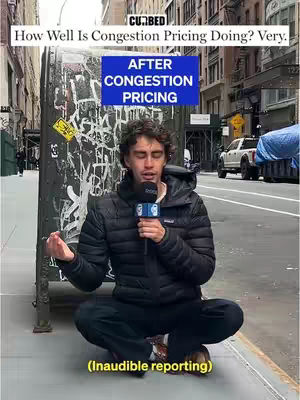 A man sits cross-legged on a city sidewalk, holding a microphone and speaking. Behind him is a weathered metal structure with graffiti, and a sign above him reads "AFTER CONGESTION PRICING". The background includes buildings and a street with vehicles.