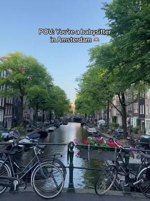 A picturesque view of a canal in Amsterdam, with bicycles parked on a bridge. Buildings line both sides of the canal, and green trees provide a natural frame.