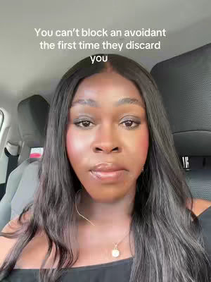 A selfie-style shot from inside a car. The main subject is a woman with dark, wavy hair and a serious expression. She's wearing a black top, and a delicate gold necklace with a pendant is visible. The background shows car headrests. Text overlayed on the image reads 'You can't block an avoidant the first time they discard you.'