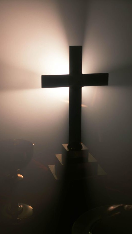 A silhouette of a cross is illuminated by bright light emanating from behind it. The cross is dark, with a long vertical beam and a shorter horizontal beam. The light creates a halo effect and visible beams. Dark vessels can be seen in the foreground, adding to the somber atmosphere.