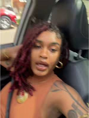 A woman with auburn hair is partially visible in the driver's seat of a car. She has a warm complexion, a nose ring, and gold hoop earrings. She's wearing a tan, sleeveless top and has a gold necklace. Tattoos are visible on her left arm. The background shows a blurry street scene with a red car. The focus is on the woman's face, with the car interior and street creating context.