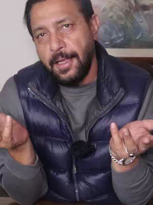 A middle-aged man with a beard and short dark hair is seated in front of a neutral background. He is wearing a gray long-sleeved shirt under a dark blue puffy vest. He has his hands up in a conversational gesture.