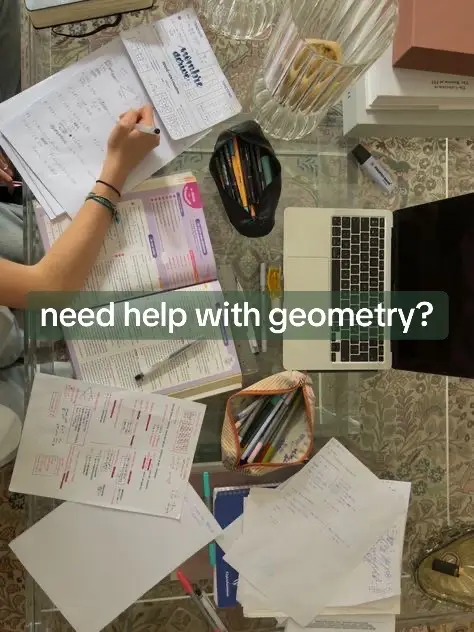 An overhead shot of a desk covered in school supplies and work. A laptop, books, stationary, and papers with notes are scattered around a glass table. A hand is writing on a paper.