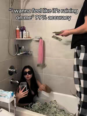 A woman in sunglasses sits in a bathtub filled with money, taking a selfie with her phone. Another person is seen in the background, throwing more cash in her direction. The scene is set in a bathroom with a shower head, toiletries, and a towel rack visible.